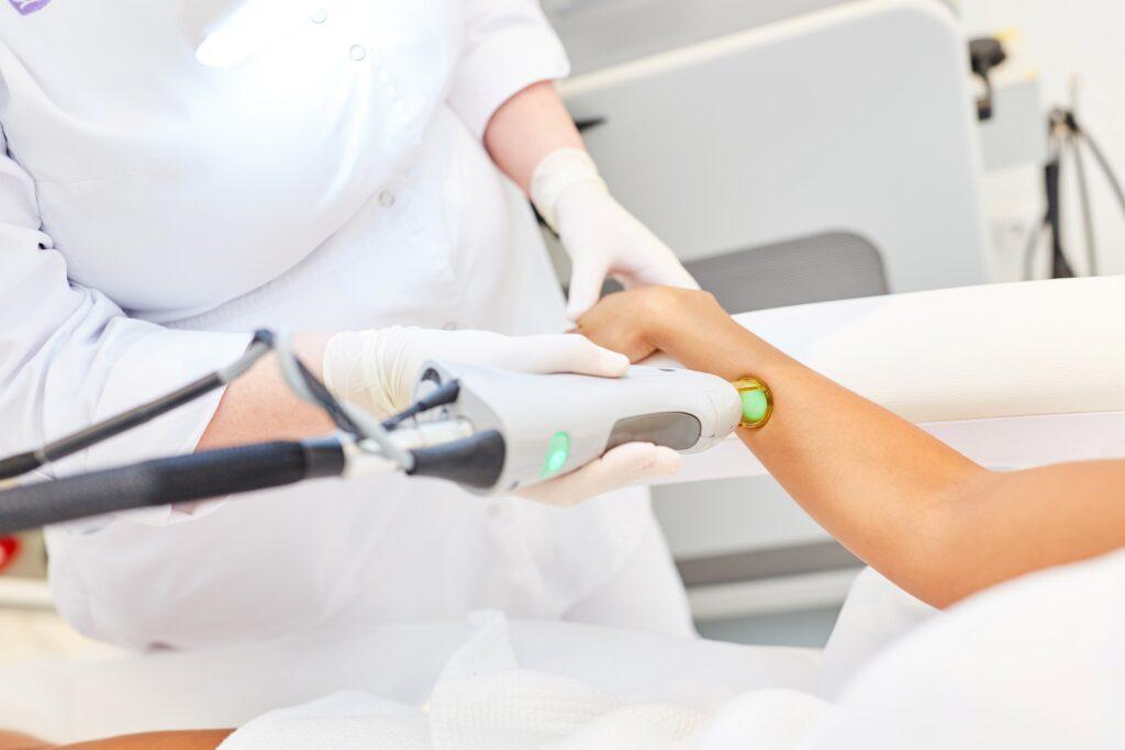 Eine Kosmetikerin führt eine Laserhaarentfernung am Unterarm einer Person durch. Beide tragen Schutzkleidung, während die Behandlung mit einem Laserapparat erfolgt.
