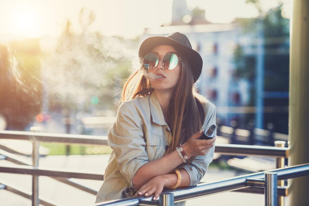 Eine junge Frau mit Sonnenbrille und Hut genießt einen entspannten Moment im Freien, während sie eine E-Zigarette mit 187 Pods verwendet. Das Bild vermittelt modernen Genuss und bewusstes Vaping als Alternative zum Rauchen.