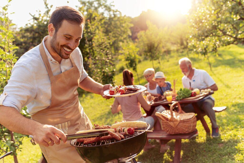 Ein Mann mit einer Schürze steht lachend am Grill und serviert gegrilltes Essen. Auf dem Grill liegen Maiskolben, Würstchen und Fleischspieße. Im Hintergrund sitzt eine Familie an einem Picknicktisch, umgeben von Bäumen, während die Sonne durch das Laub scheint und eine warme, entspannte Atmosphäre schafft.